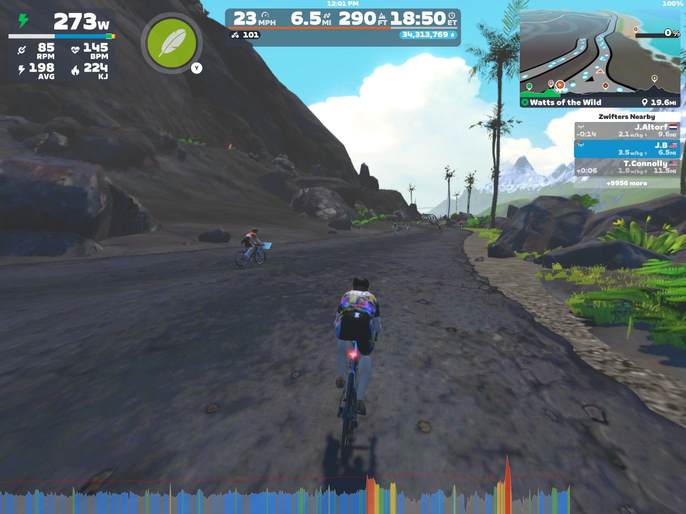 A cyclist rides on a rugged path in a virtual landscape, surrounded by rocks and greenery. The display shows performance stats, including speed and distance, along with a mini-map indicating the route. Additional cyclists are visible in the background, and mountains can be seen in the distance under a blue sky.